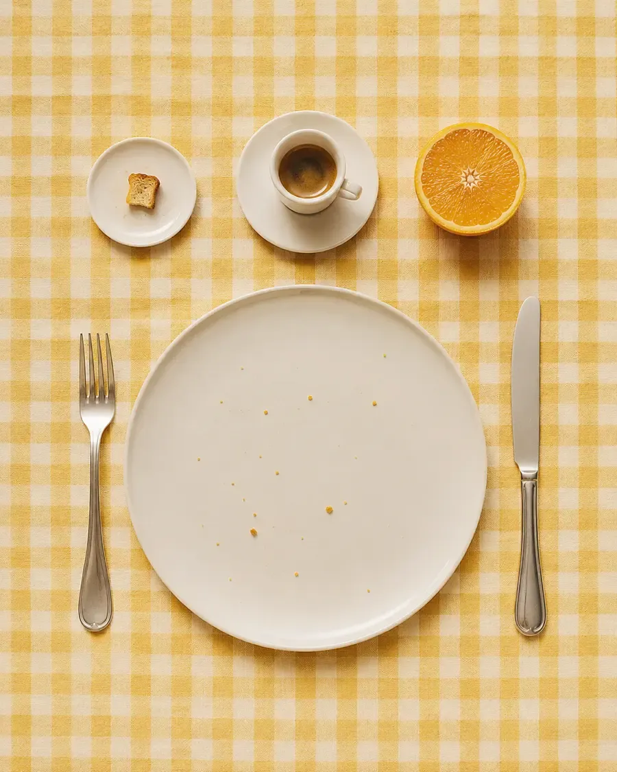 A minimalist yellow gingham table setting with a large empty white plate, a small cup of espresso, half an orange, a tiny piece of bread on a saucer, and silverware — a quiet visual of how "light" breakfasts often look | Becoming Elysian quiet luxury wellness